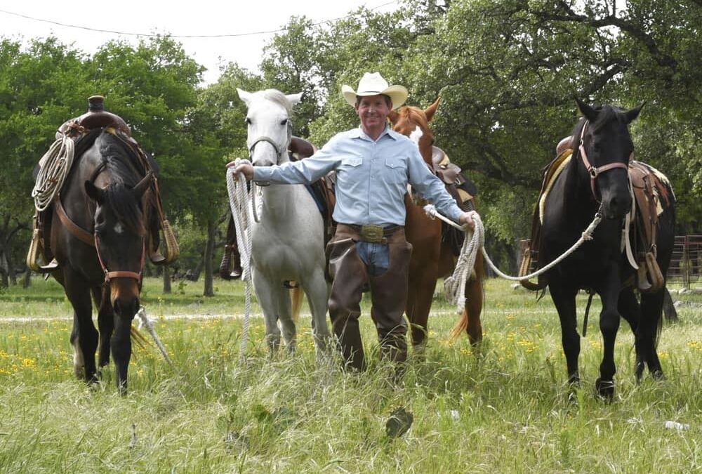 The Cowboy Entrepreneur Scott Knudsen wears a cowboy hat and Western attire as he leads four horses through a grassy field