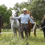 The Cowboy Entrepreneur Scott Knudsen wears a cowboy hat and Western attire as he leads four horses through a grassy field