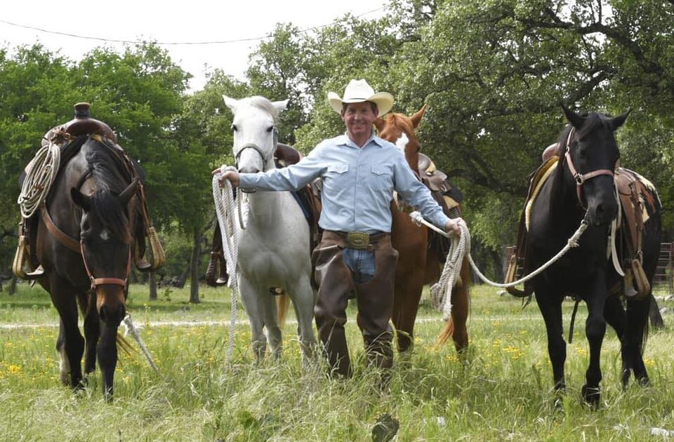 The Cowboy Entrepreneur Scott Knudsen wears a cowboy hat and Western attire as he leads four horses through a grassy field